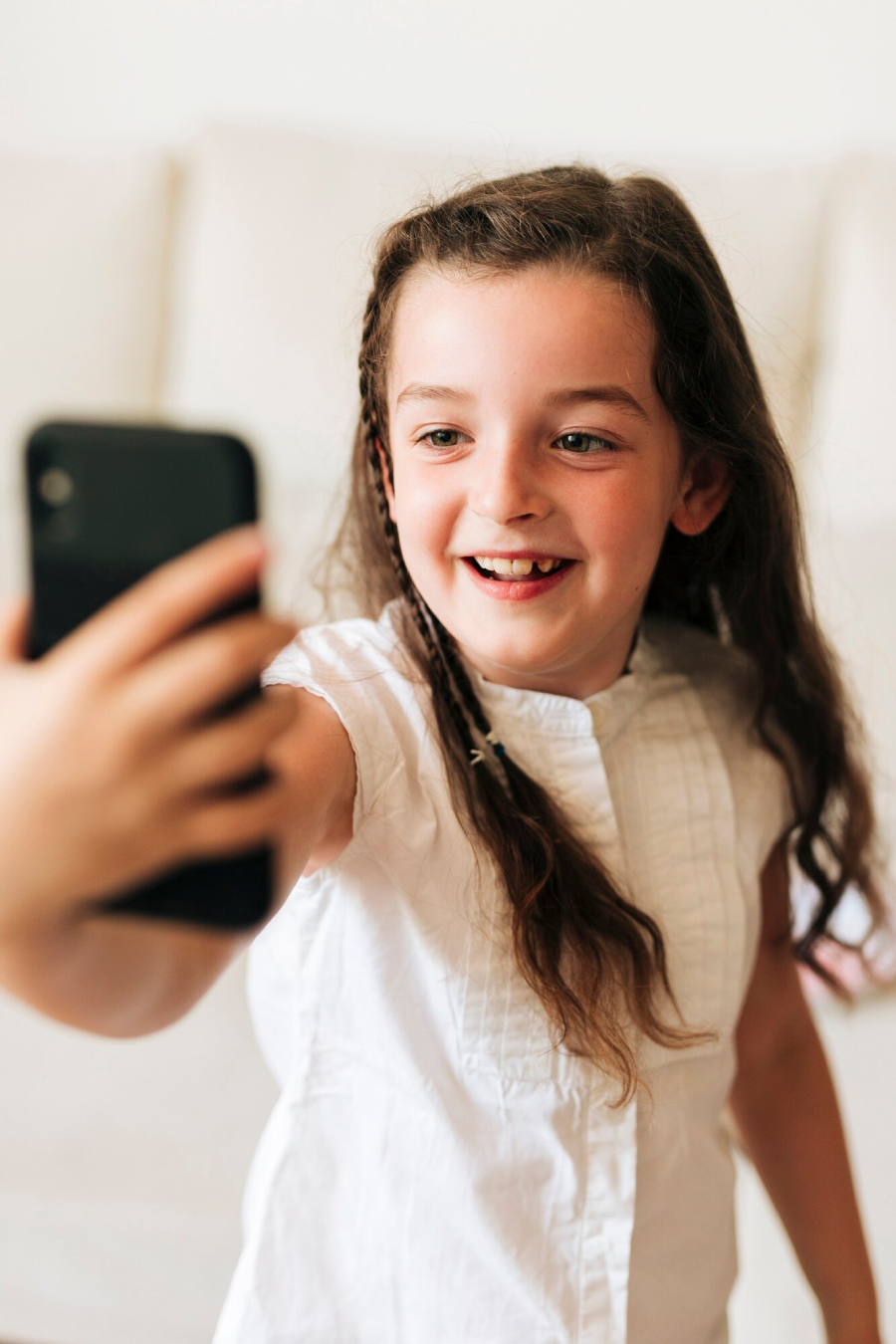 A smiling young girl with long brown hair wearing a white shirt holds up a smartphone, appearing to take a selfie indoors.