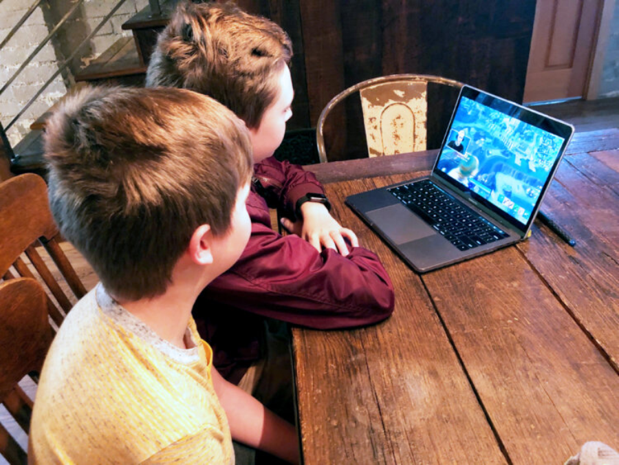 Two boys sit at a wooden table, watching something on a laptop screen together. The laptop displays a colorful scene, and both boys are focused on the screen.