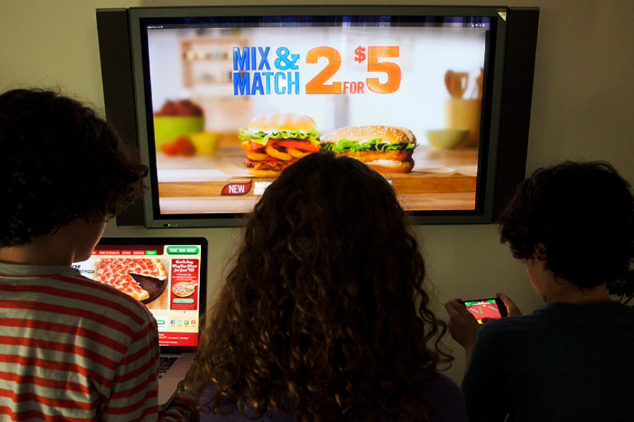 Three children face a TV displaying a sandwich ad, “Mix & Match 2 for $5.” One uses a laptop with food images, another looks at a phone, and the third watches the screen. All devices show food-related content.