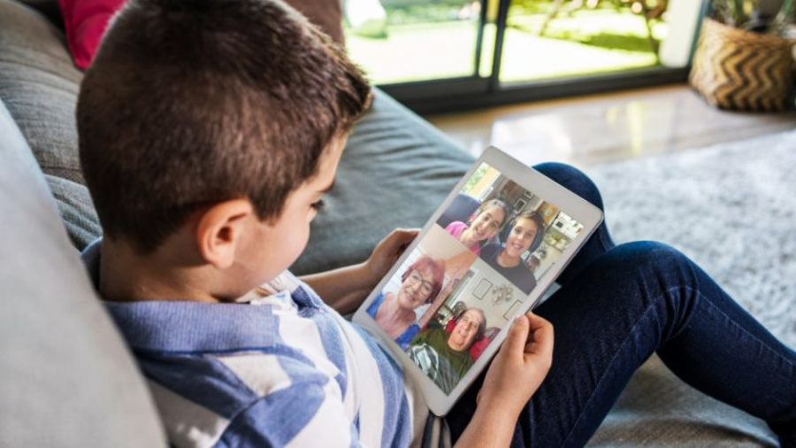 A boy sits on a couch holding a tablet, video calling with four people whose faces are visible on the screen. Sunlight comes through glass doors in the background.