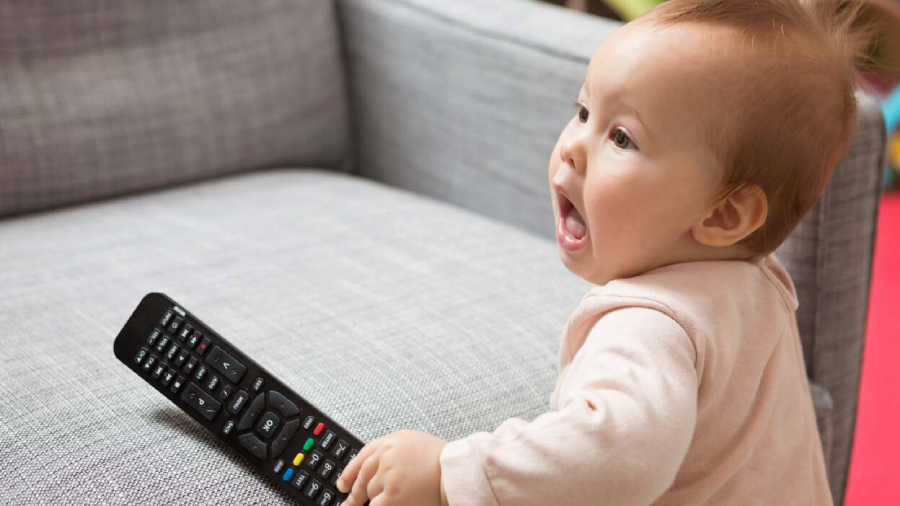 A baby with an excited expression sits on a gray couch, reaching for a black TV remote control placed on the seat beside them.
