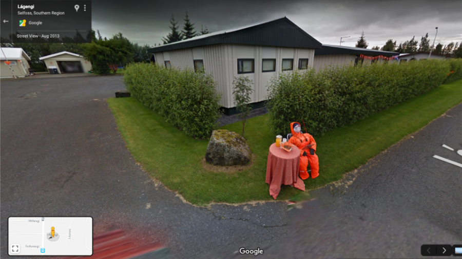 A person in an orange hazmat suit sits at a small round table with a pink tablecloth, set with cups and a teapot, on a grassy corner outside a building with light walls and a dark roof.