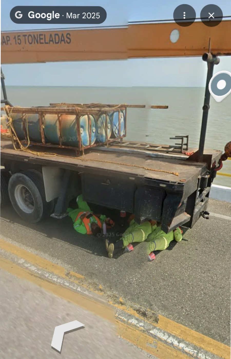 Two construction workers in green uniforms and helmets are lying under a large flatbed truck with a crane near a body of water, appearing to inspect or repair something beneath the vehicle.