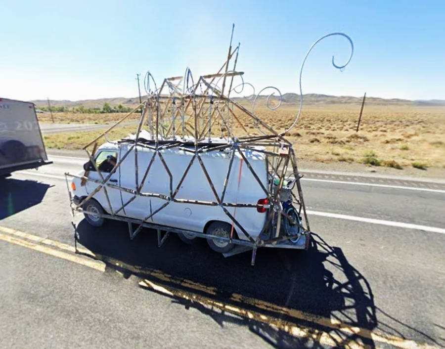 A white van is driving down a highway with an elaborate, whimsical metal sculpture frame attached to its roof and sides, extending high above and around the vehicle. Dry, grassy scenery is visible in the background.