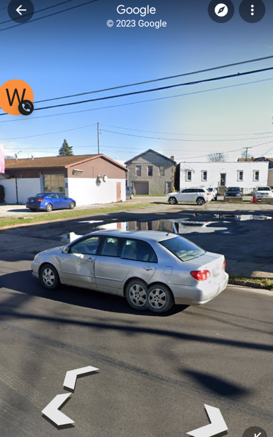 A silver sedan drives down a residential street with single-story houses and parked cars. There's a large puddle on an empty lot, and the sky is clear and sunny.