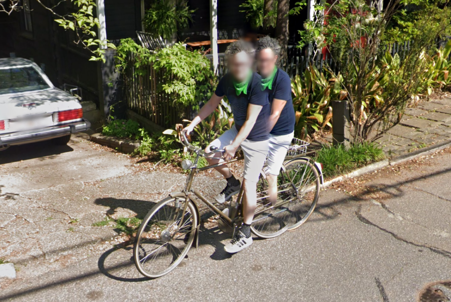 Two people wearing matching navy shirts and green scarves are riding together on a tandem bicycle along a residential street, passing by greenery and a parked white car.