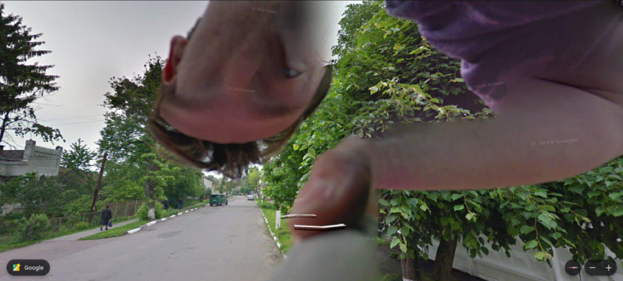 A distorted image of a person’s face and arm appears close to the camera, seemingly on a quiet street lined with green trees and a sidewalk. The Google Street View watermark is visible in the corner.