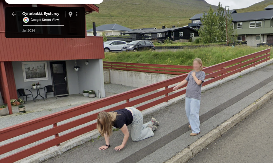 Two people are on a sidewalk beside a red fence. One is on hands and knees; the other stands nearby, appearing surprised. Houses and a grassy hill are in the background. The Google Street View label shows "Oyrarbakki, Eysturoy.