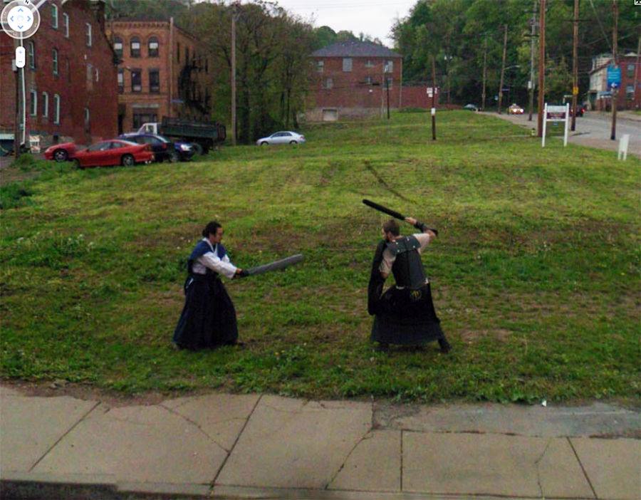 Two people in medieval-style clothing are mock fighting with foam swords on a grassy lot in an urban area, with brick buildings and parked cars in the background.