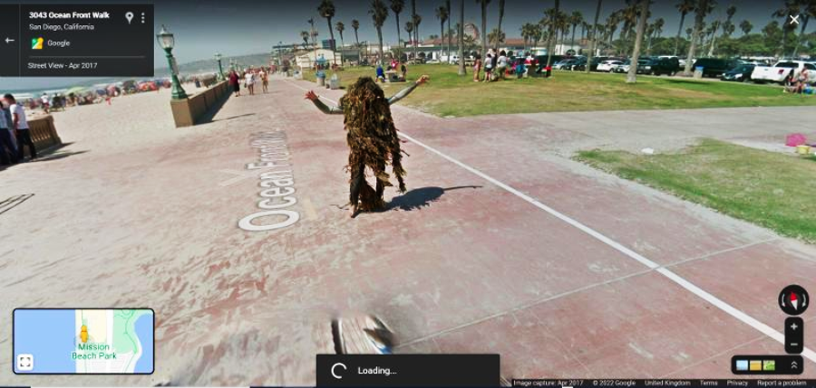 A person in a costume made of brown, tattered material stands with arms raised on a seaside walkway in San Diego, near grass, palm trees, and the beach. People are in the background enjoying the sunny day.