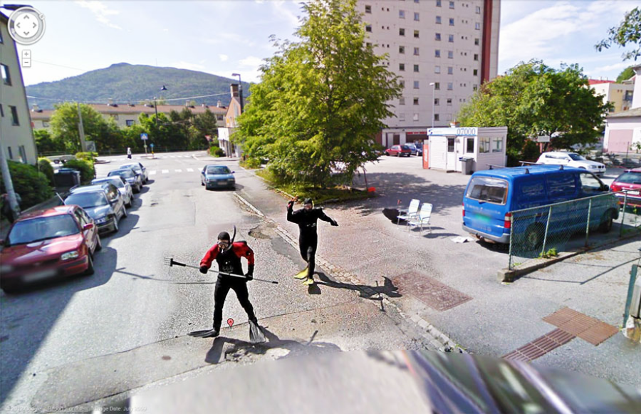Two people in roller ski gear pose playfully in the middle of a quiet street next to parked cars, with buildings, trees, and a mountain in the background on a sunny day.