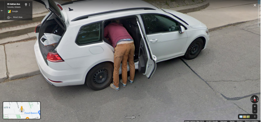 A person wearing a maroon jacket and tan pants is bent over, reaching inside the back seat of a white station wagon parked on a street. The car's rear door and trunk are open. A city sidewalk and curb are visible.