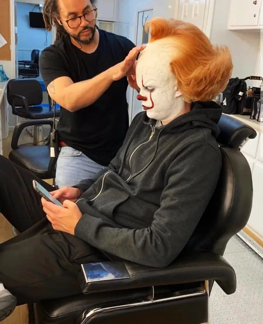 A person dressed as Pennywise the clown from "It" sits in a makeup chair, using a phone, while a makeup artist styles their orange wig in a dressing room.
