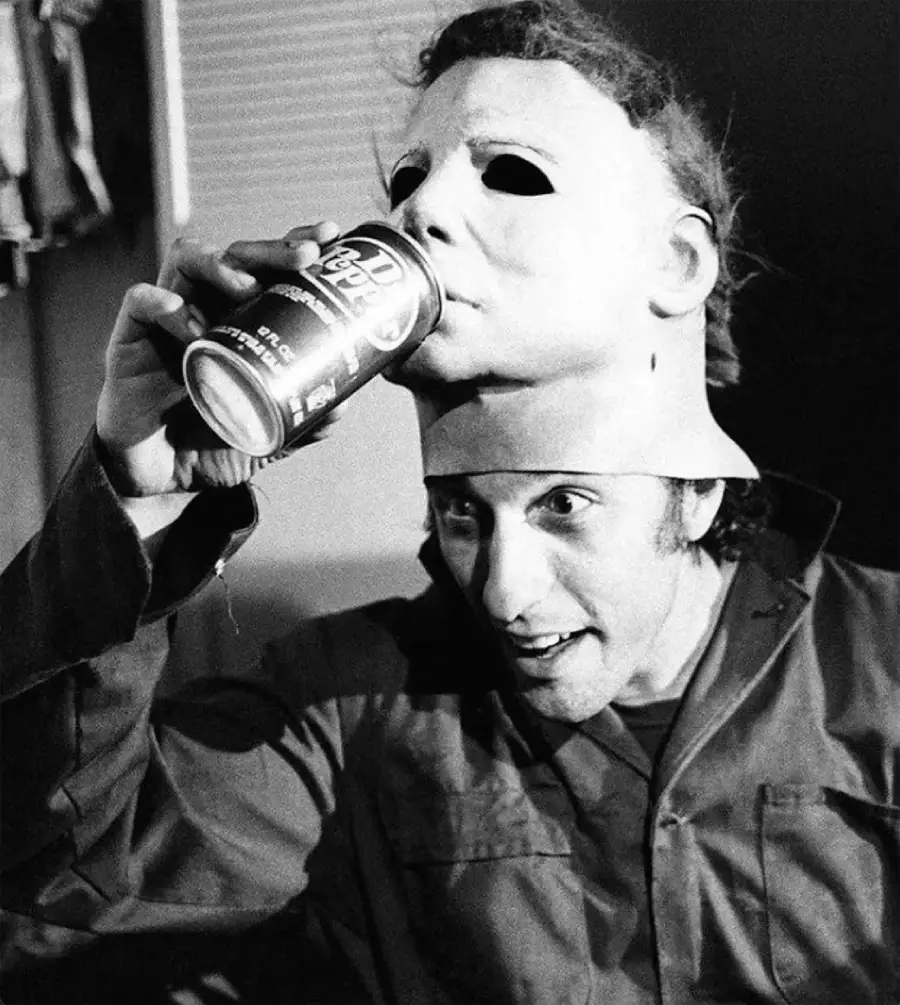 A person in a jumpsuit lifts a Michael Myers mask above their head and drinks from a soda can, with the mask resting on top of their head. The photo is in black and white.