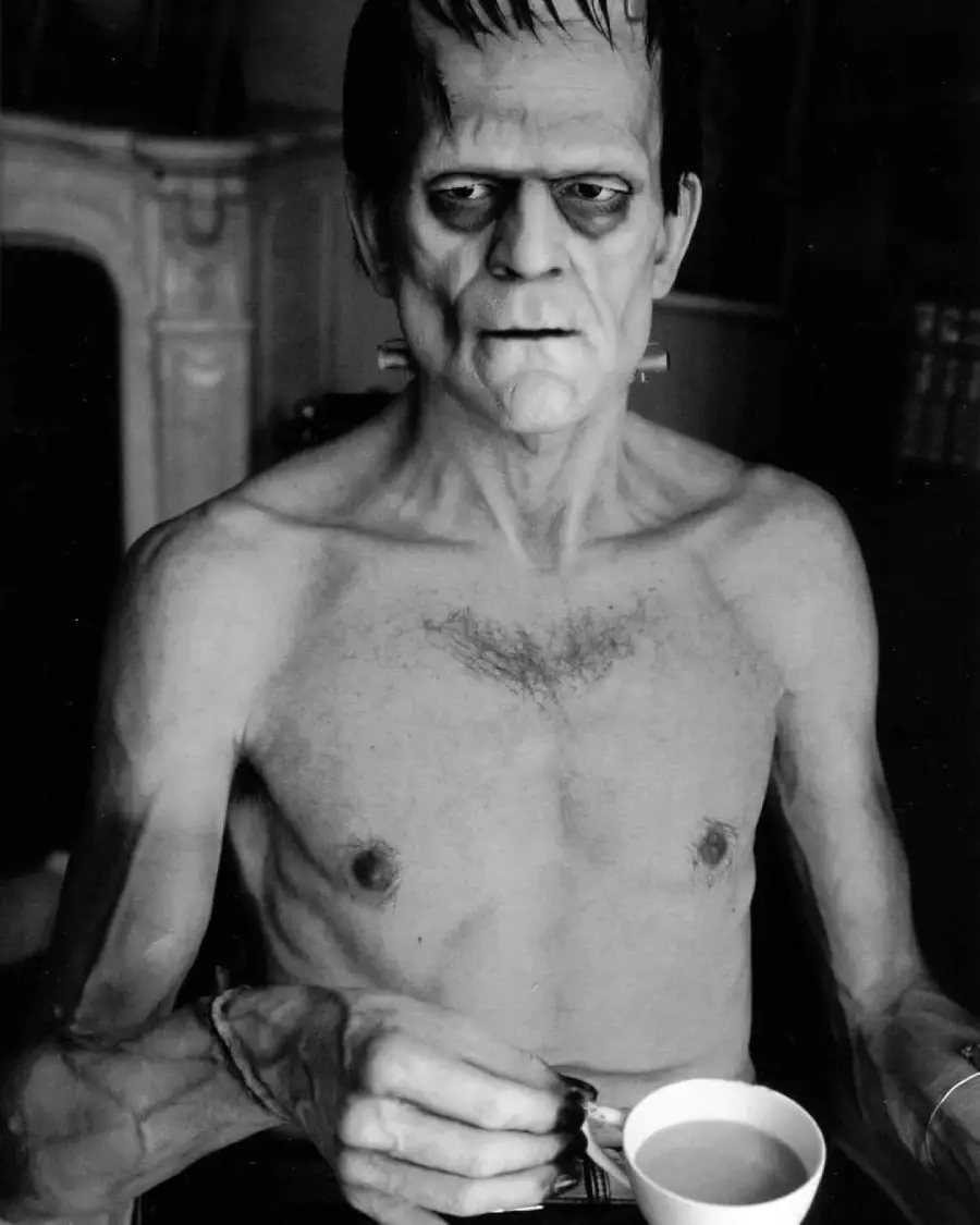 A shirtless person in Frankenstein's monster makeup holds a teacup and saucer, looking off to the side with a serious expression. The photo is black and white and taken indoors.