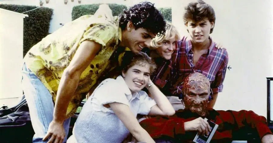 Five people pose together outdoors, four smiling teens and one adult in detailed monster makeup and costume, resembling Freddy Krueger from "A Nightmare on Elm Street." They appear to be enjoying a break during filming.