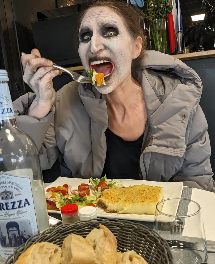 A person with dramatic white face makeup and dark eye circles, dressed in a light gray coat, eats salad at a restaurant table set with fish, bread, and a bottle of mineral water.