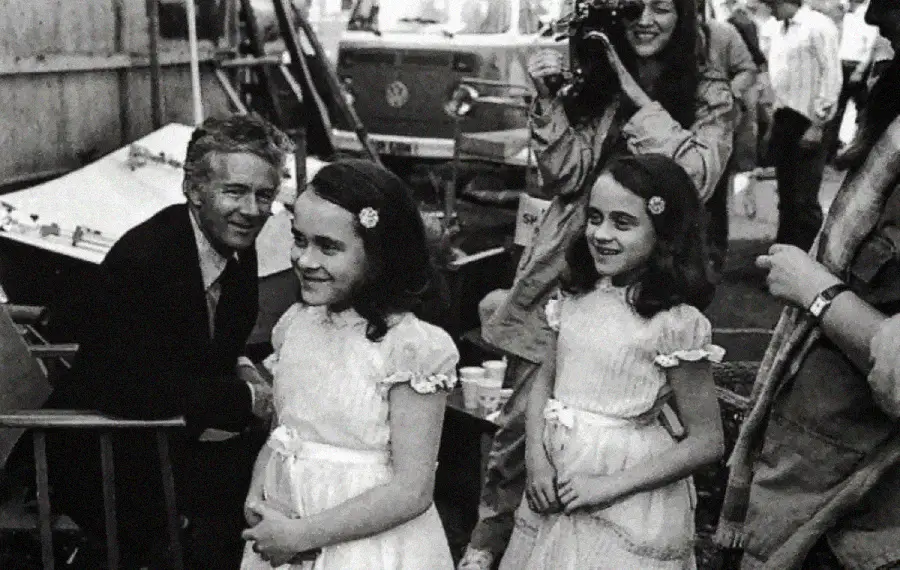 Two young girls in matching dresses stand side by side, smiling. A man crouches beside them, also smiling. A woman behind them holds a camera, and other people and equipment are visible in the background.