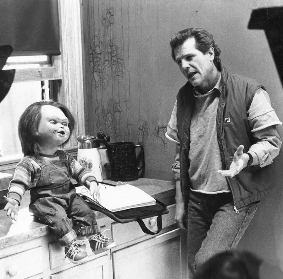 A man gestures while talking to a puppet that resembles Chucky, the doll from the Child’s Play horror movies, sitting on a table next to an open script in a behind-the-scenes setting.