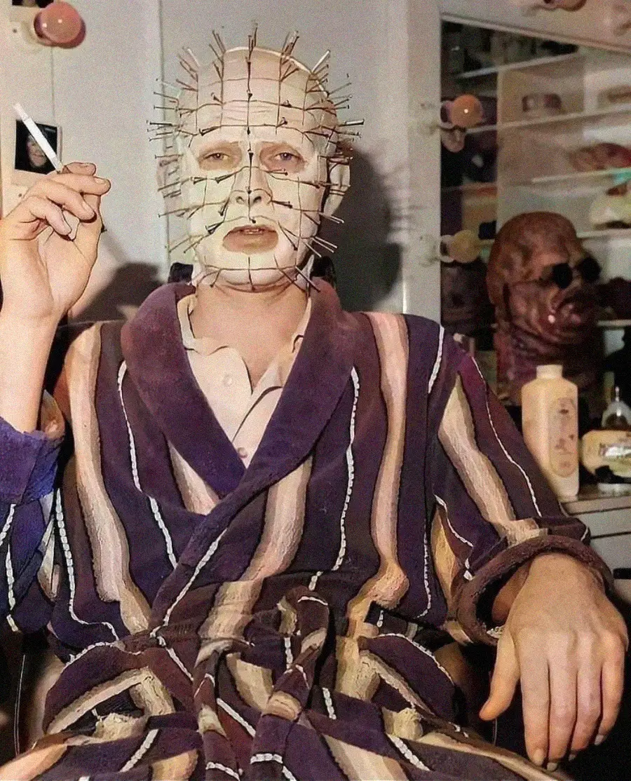 A person wearing a striped robe and a mask covered with pins sits holding a cigarette. The background includes shelves with various objects, including a bust and bottles.