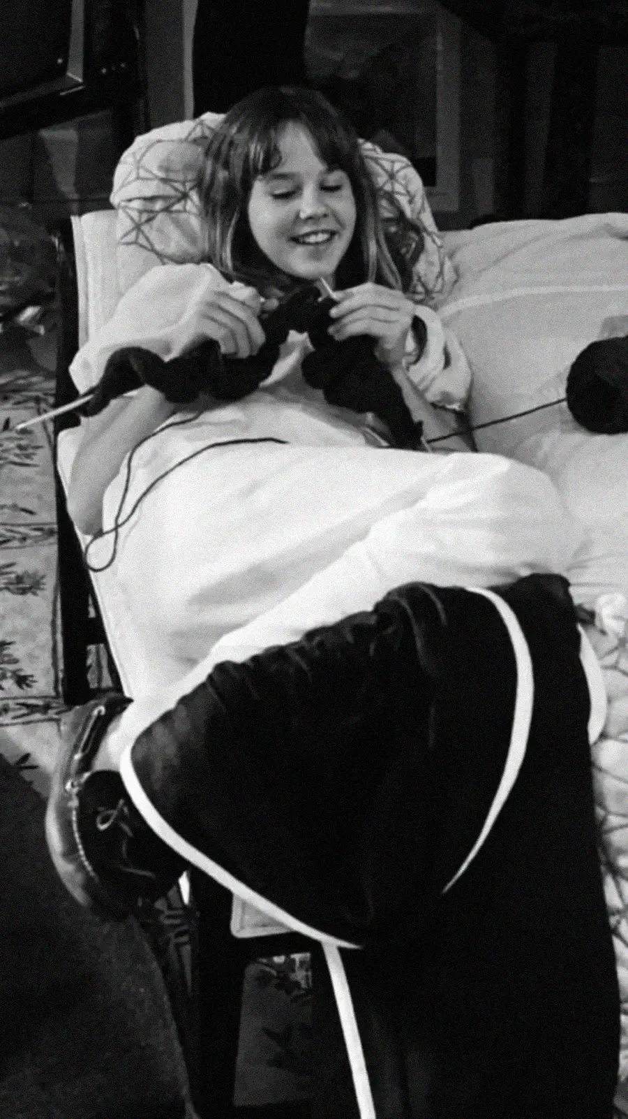 A young girl lounges comfortably on a couch with a blanket, smiling as she knits with dark yarn. She appears relaxed and content, with her legs propped up and crossed. The photo is in black and white.