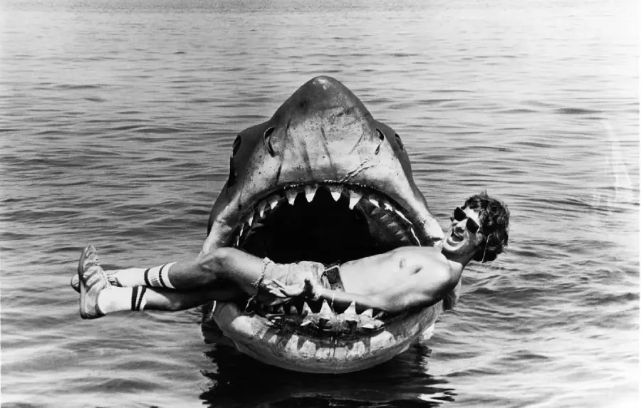 A man wearing sunglasses and shorts playfully poses inside the open mouth of a large, fake shark in the water, appearing as if he is being eaten.