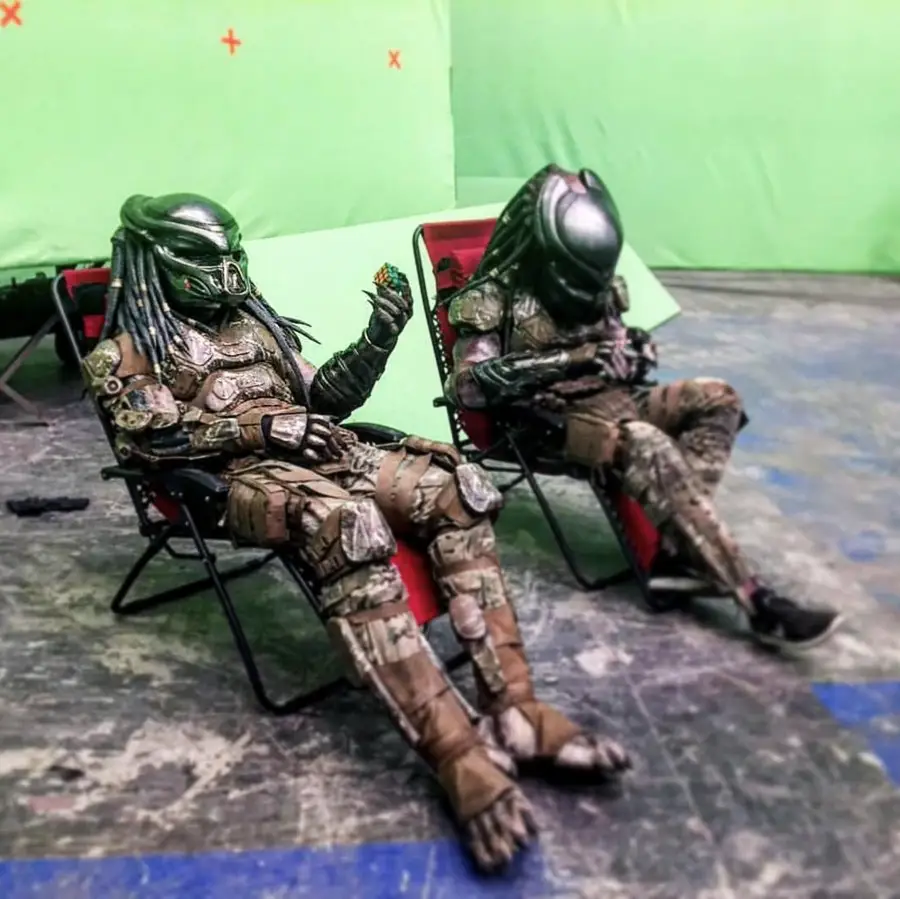 Two people in detailed Predator costumes sit on red folding chairs on a studio set with a green screen background. One looks at a Rubik's Cube while the other is looking down, possibly at a phone or similar object.