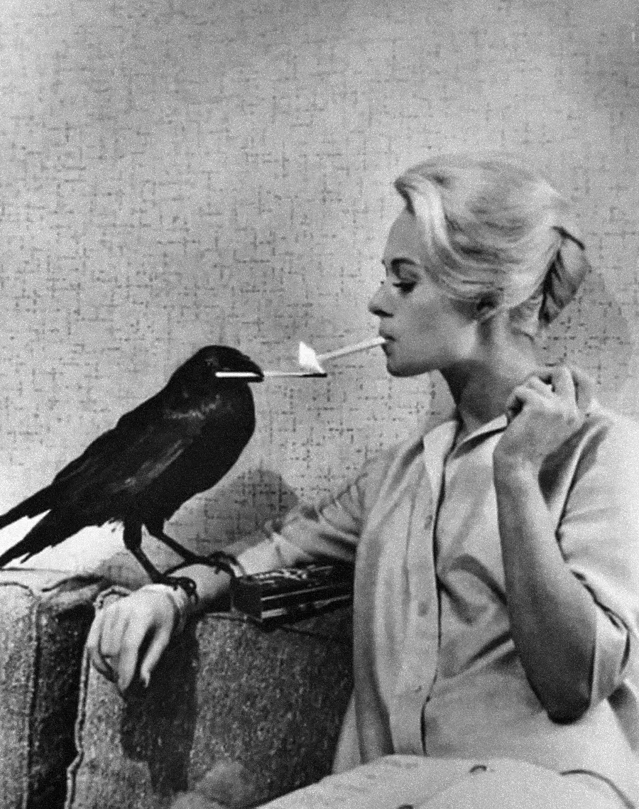 A woman with light hair holds a cigarette in her mouth while a crow sits on her arm, appearing to offer her a match to light the cigarette. The scene is in black and white.