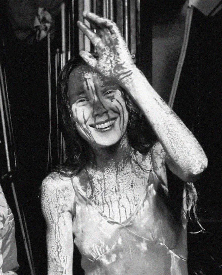 A woman in a white dress, covered in a dark liquid, smiles and waves at the camera. The image is black and white, and her hair and face are also splattered with the liquid.
