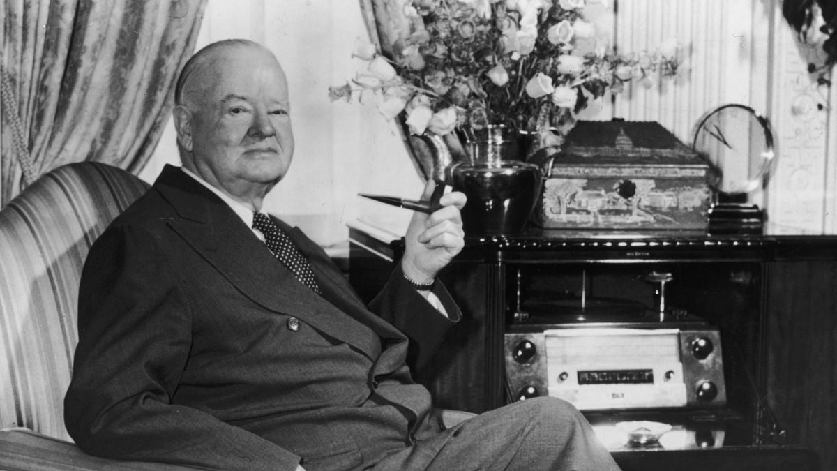 Elderly man in a suit sits in an armchair, holding a pipe and smiling. A vase of flowers, ornate box, and old-fashioned radio are on the table behind him. Heavy drapes and a decorative wall are in the background.
