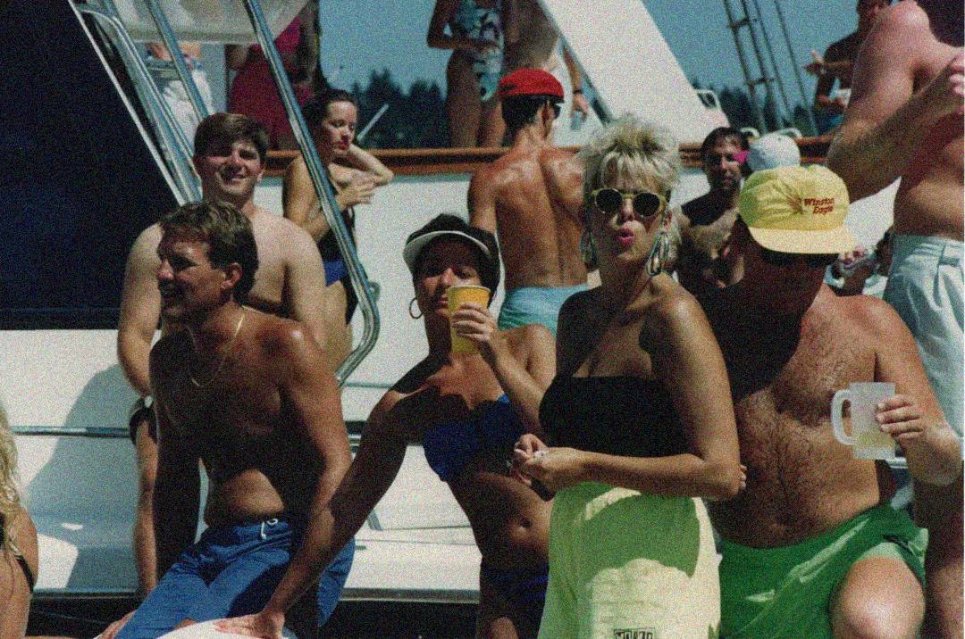 A group of people in swimsuits and summer clothes enjoy drinks and socialize on a boat under the sun, with more boats visible in the background.
