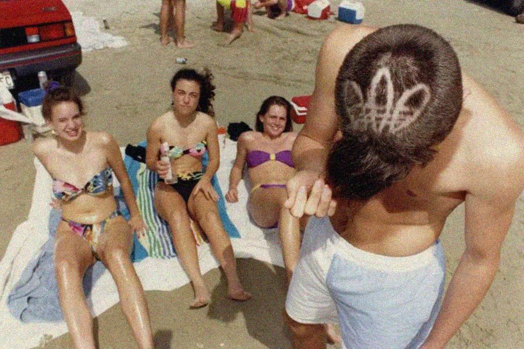 A group of three women in swimsuits sit on towels at the beach while a man stands in front of them, showing a haircut with the Adidas logo shaved onto the back of his head. There are beach bags and coolers in the background.