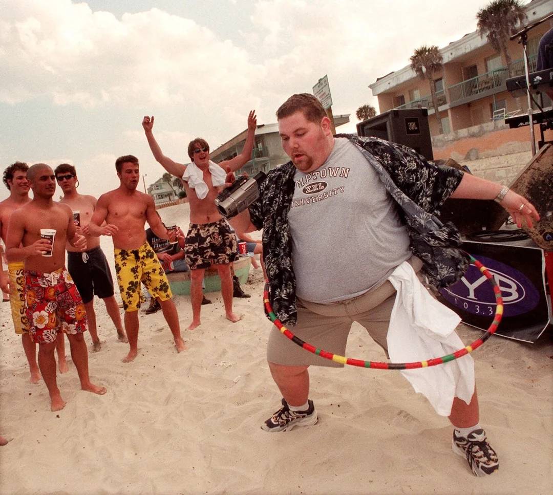 A man in a patterned shirt and gray T-shirt hula-hoops on a sandy beach while a group of young men in swim trunks cheer him on. Buildings and palm trees are visible in the background.