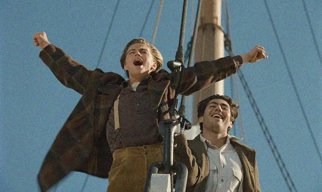 Two men stand at the bow of a ship; one has his arms outstretched joyfully while the other stands behind him, smiling, with a mast and blue sky in the background.