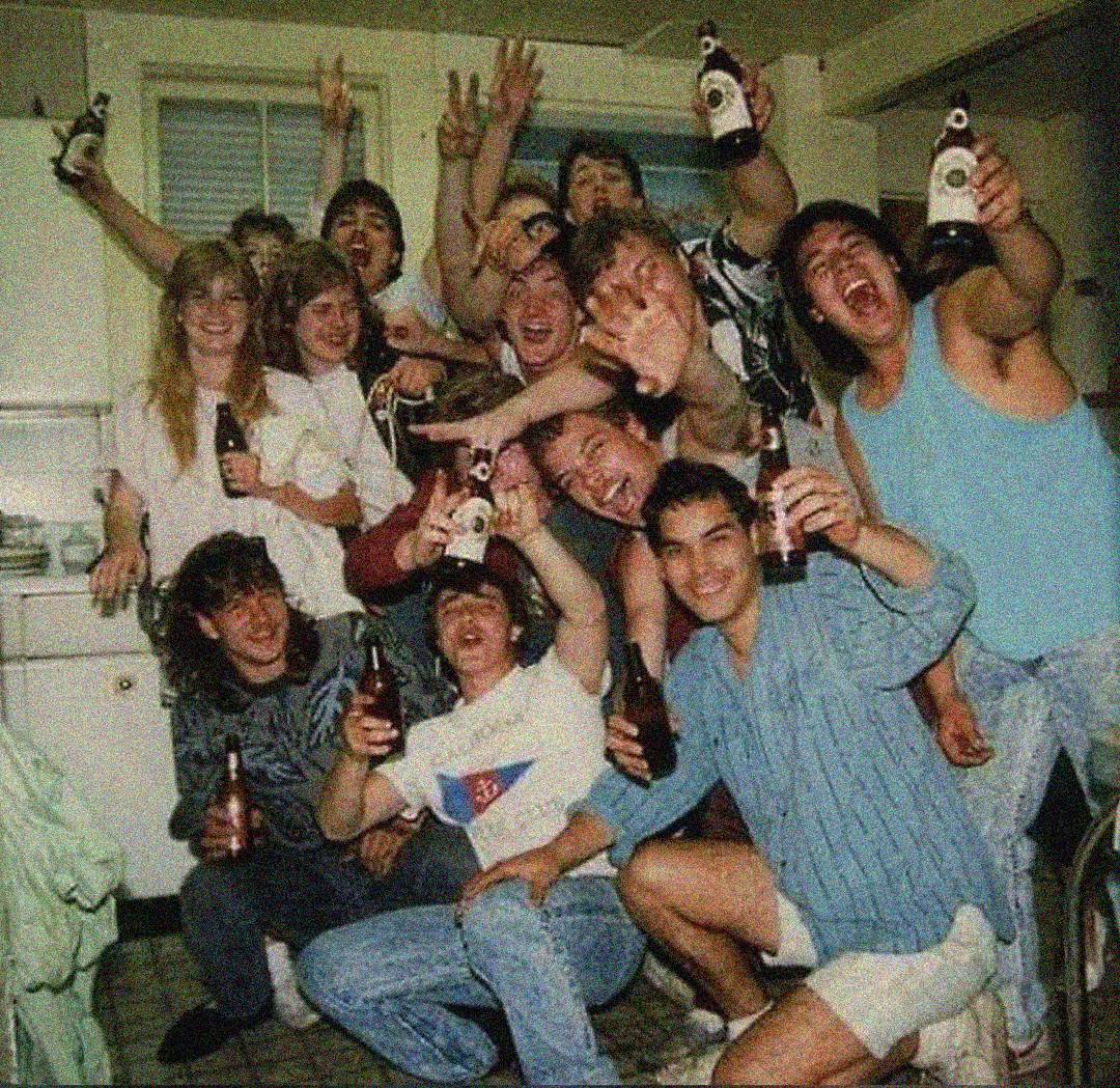 A group of young adults in a kitchen, smiling, laughing, and holding beer bottles, appearing to celebrate and pose enthusiastically for the camera.