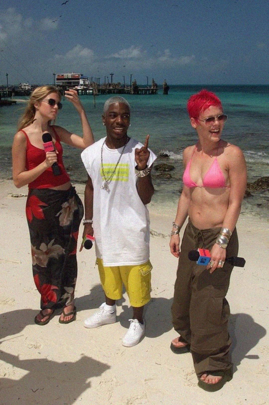 Three people stand on a beach holding microphones. One wears a pink bikini top and brown pants, another wears yellow shorts and a white t-shirt, and the third wears a red and black floral outfit. The ocean and pier are in the background.
