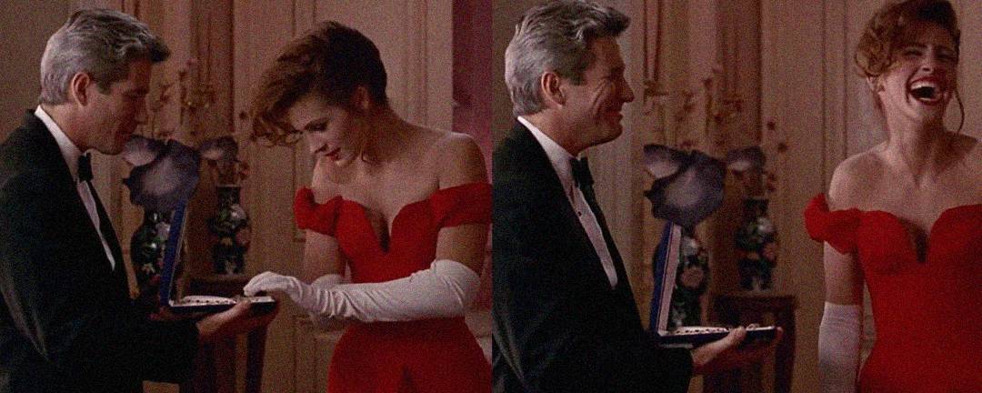 A man in a tuxedo presents a necklace to a woman in a red off-shoulder gown and white gloves; she laughs joyfully as he playfully snaps the box shut.