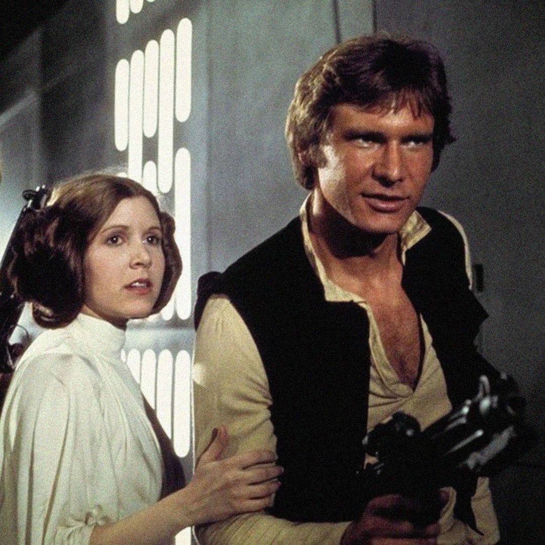 A woman with brown hair in side buns stands beside a man with brown hair, wearing a vest and holding a blaster, in a sci-fi setting with a glowing panel in the background.
