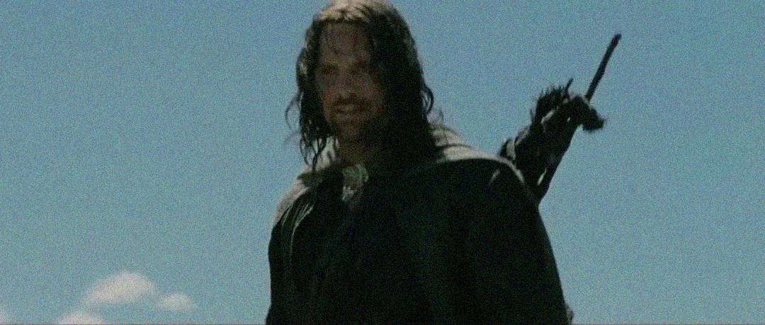 A man with long dark hair and a rugged appearance stands outdoors under a clear blue sky, wearing a dark cloak and carrying a sword on his back.