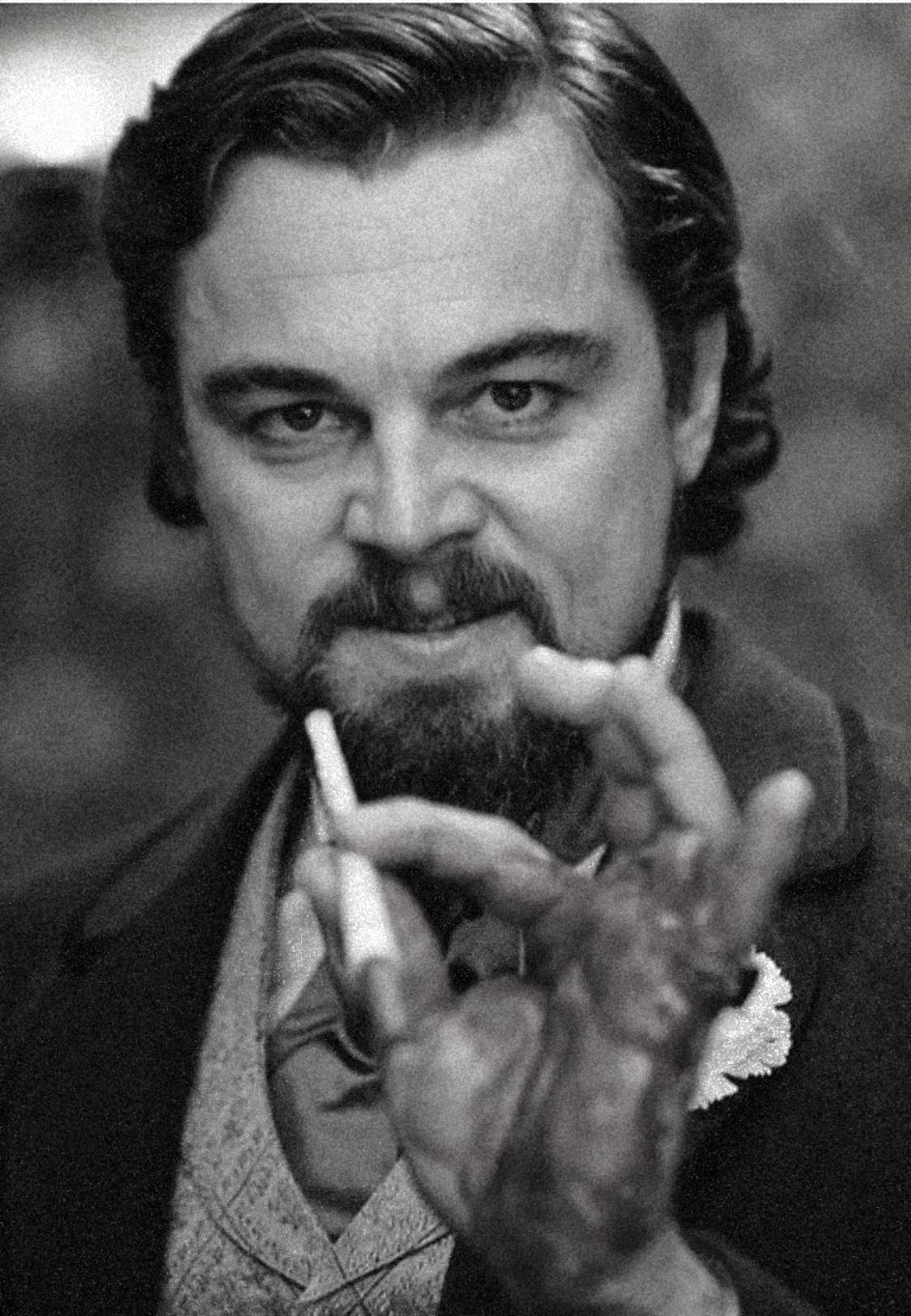 A man with a styled beard and mustache holds a cigarette between his fingers, which appear bloodied. He wears a suit with a boutonnière, gazing intently at the camera in a black-and-white photo.
