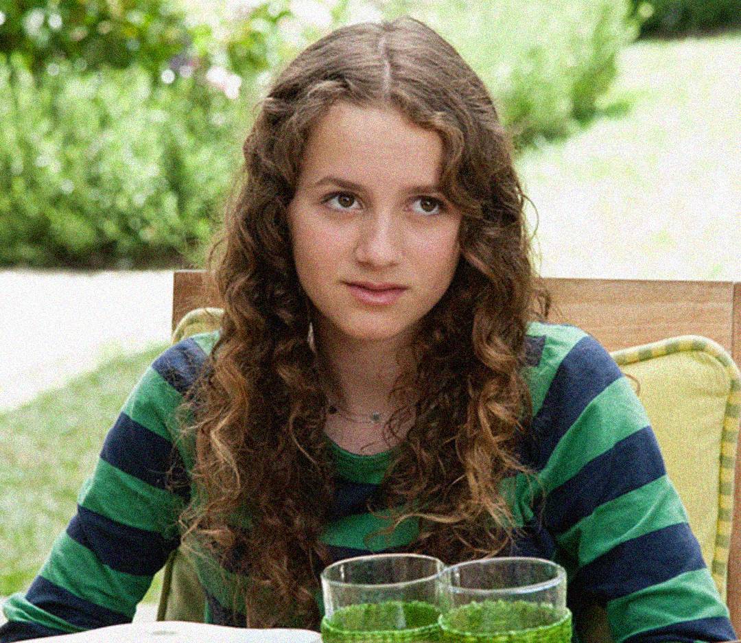 A young person with long curly brown hair wearing a green and navy striped shirt sits outdoors at a table with two glasses of a green drink in front of them. The background is bright and blurred with greenery.