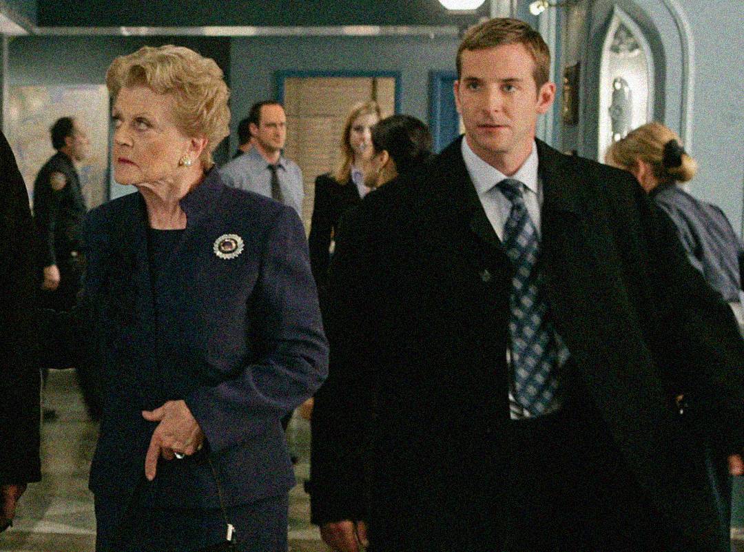 An older woman in a navy suit and a younger man in a black coat and tie walk through a busy hallway with several people in the background. Both appear focused and serious.