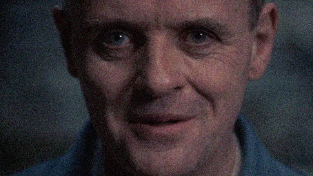 A close-up of a middle-aged man with light skin, blue eyes, and a slight smile, gazing directly at the camera. The background is dimly lit and out of focus.