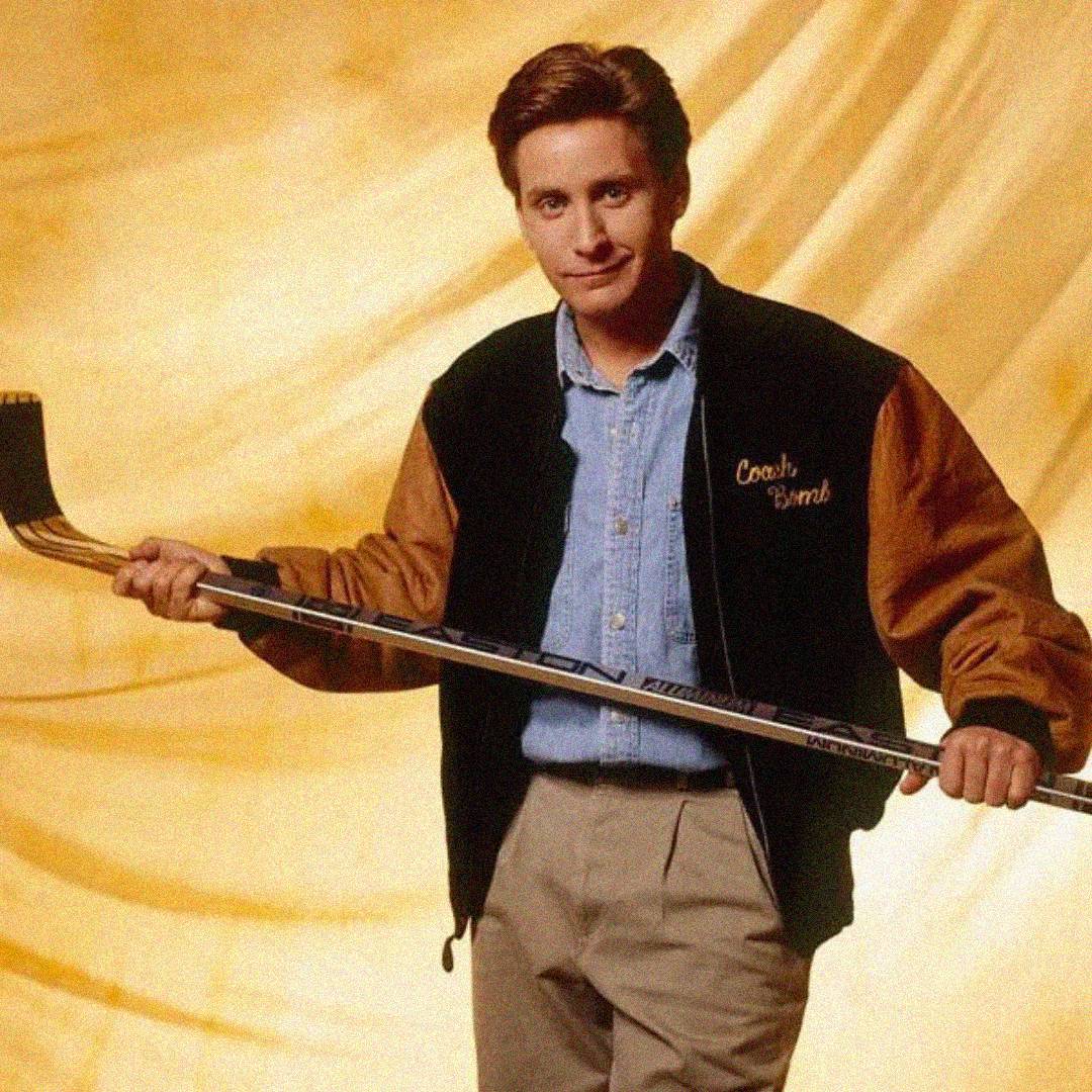 A man with short brown hair stands holding a hockey stick horizontally. He wears a blue shirt, tan pants, and a black and brown jacket with "Coach Bombay" embroidered on it, against a golden, softly lit background.
