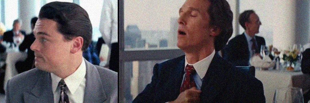 Two men in suits sit at a restaurant table. The man on the left looks to his right with a serious face, while the man on the right leans back, eyes closed, hitting his chest with his fist as if humming or making a sound.