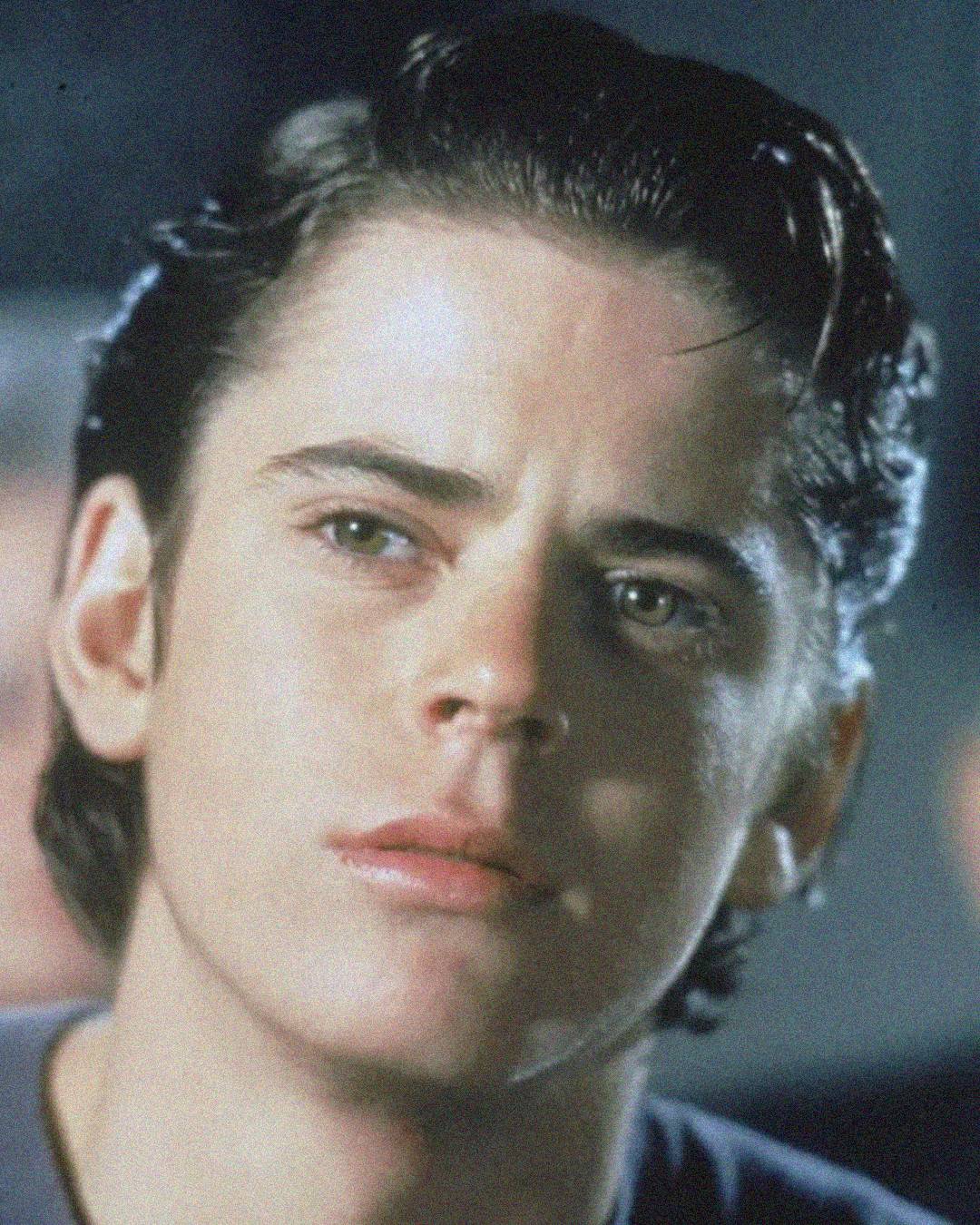 A young person with short, dark, slicked-back hair and light eyes looks slightly to the side with a serious expression on their face. The background is out of focus.