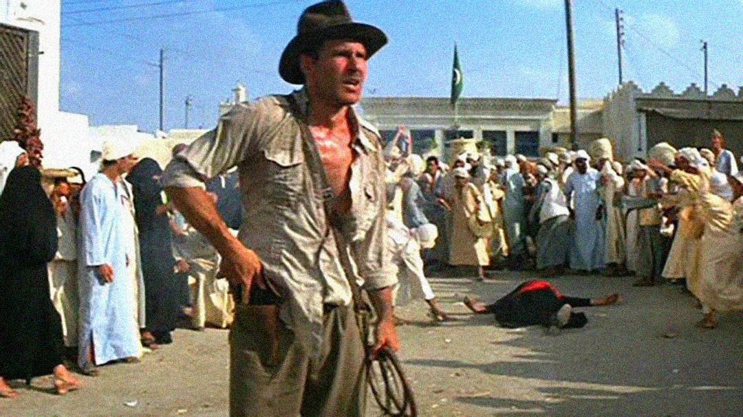 A man in a fedora hat and dusty shirt stands holding a whip in a crowded marketplace, with onlookers around him and a person lying on the ground in the background.