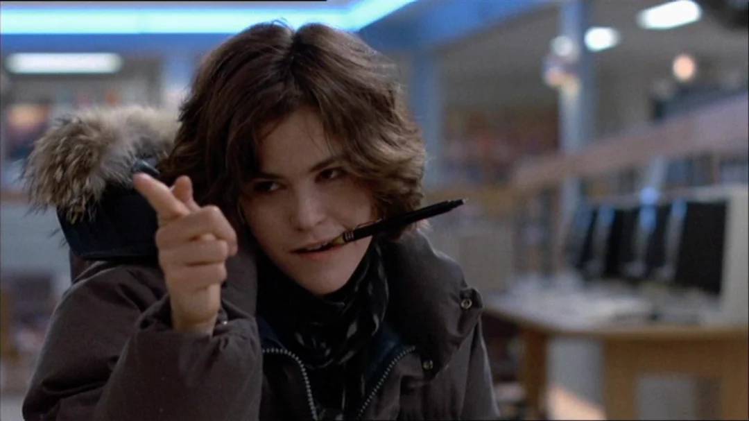 A young woman with short brown hair sits indoors wearing a dark coat with a fur-lined hood. She holds a pen in her mouth and points her fingers playfully, with computers visible in the background.
