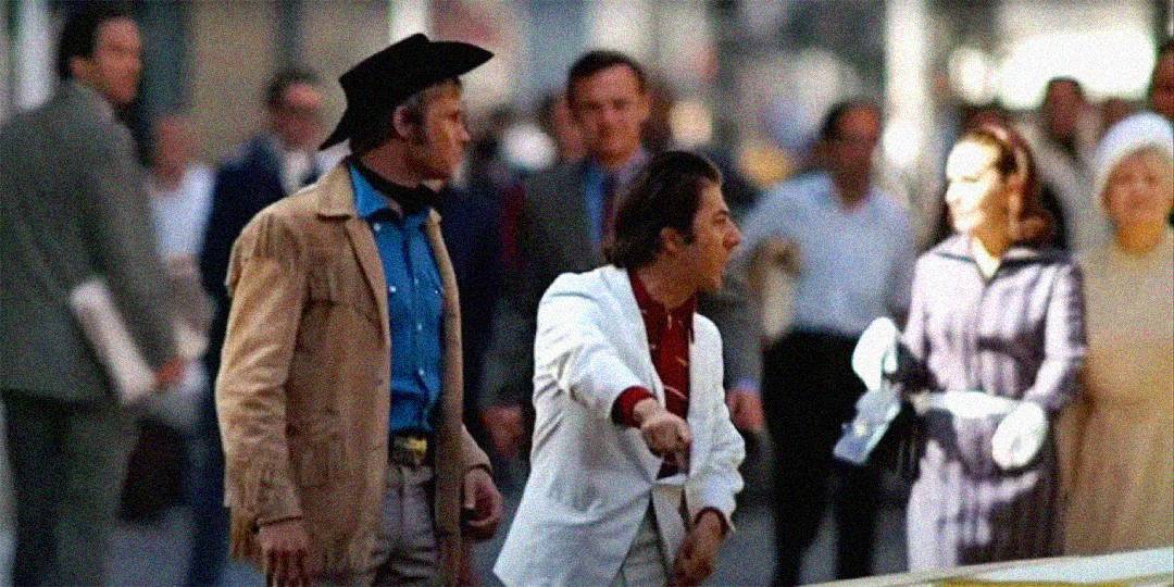 Two men stand on a busy city street; one wears a cowboy hat and tan jacket, and the other wears a white jacket and gestures outward. Pedestrians and blurred figures move around them in the background.