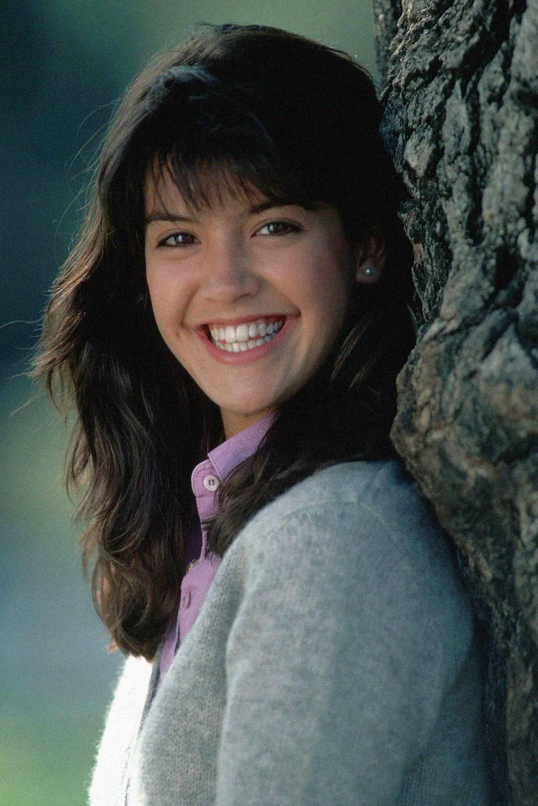 A young woman with long dark hair and bangs smiles brightly next to a tree, wearing a light gray sweater over a lavender collared shirt, with a blurred green background.